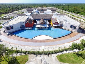 A large swimming pool with a surrounding walkway, multiple buildings with flat roofs, and a clear sky. - Atabey Luxury Paradise (Punta Cana)