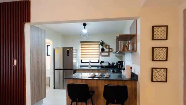 Eine moderne Küche mit Kochinsel, zwei Barhocker, einem Kühlschrank und einem Fenster mit Jalousien.