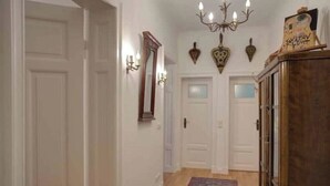 Un couloir aux murs blancs, doté d’un plancher en bois et orné d’un tapis à motifs. On y trouve deux portes, un lustre et des appliques murales.