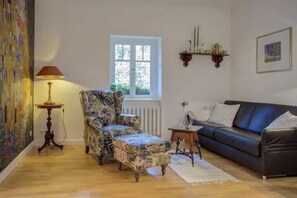 A living room with a patterned armchair, a sofa, a small table with a lamp, and a window with a view of greenery. - Strandvilla Hubertus - H3 - Gustav Klimt (Ostseebad Boltenhagen)