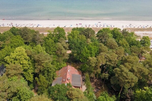 An aerial view of a coastal area with a beach, dense forest, and a house surrounded by trees.
