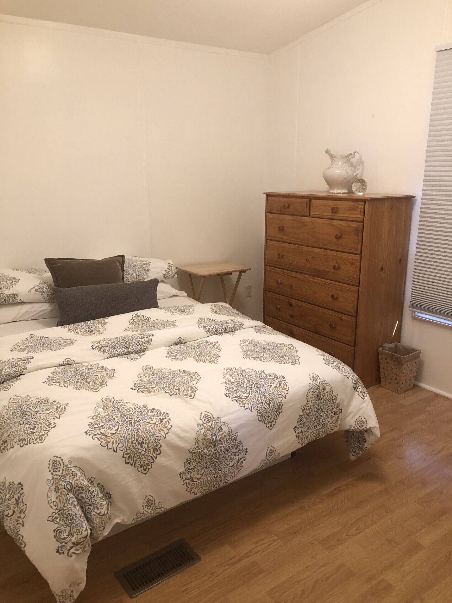 Ein Schlafzimmer mit einem Bett, einer Holzkommode, einem kleinen Tisch und einem Fenster mit Jalousien.