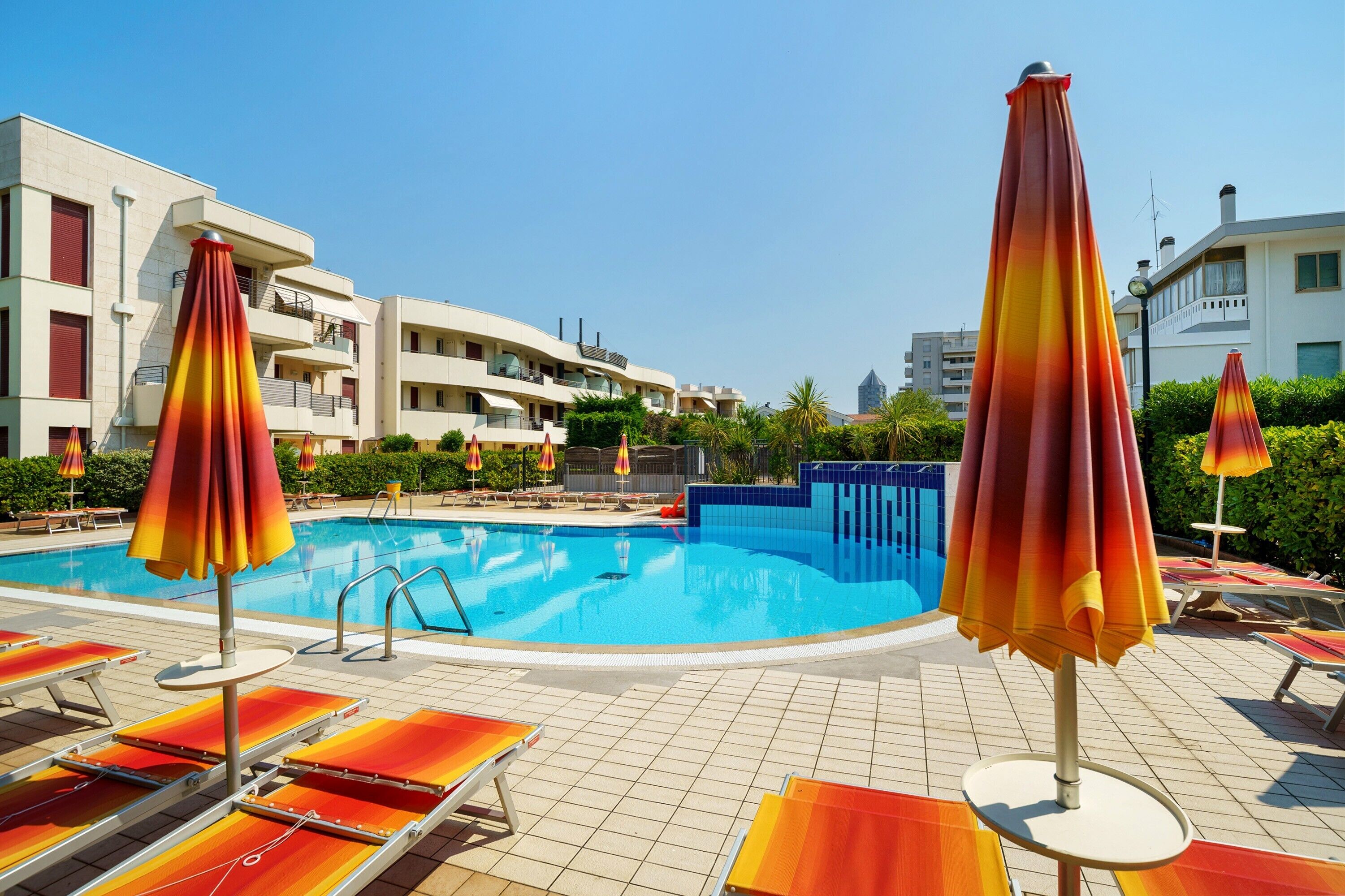 A swimming pool with sun loungers and umbrellas, surrounded by buildings.