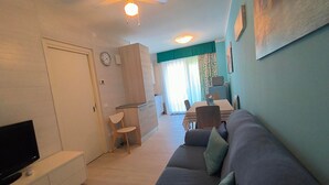Una sala de estar con sofá, televisor, zona de comedor con mesa y sillas, ventilador de techo y un reloj en la pared.