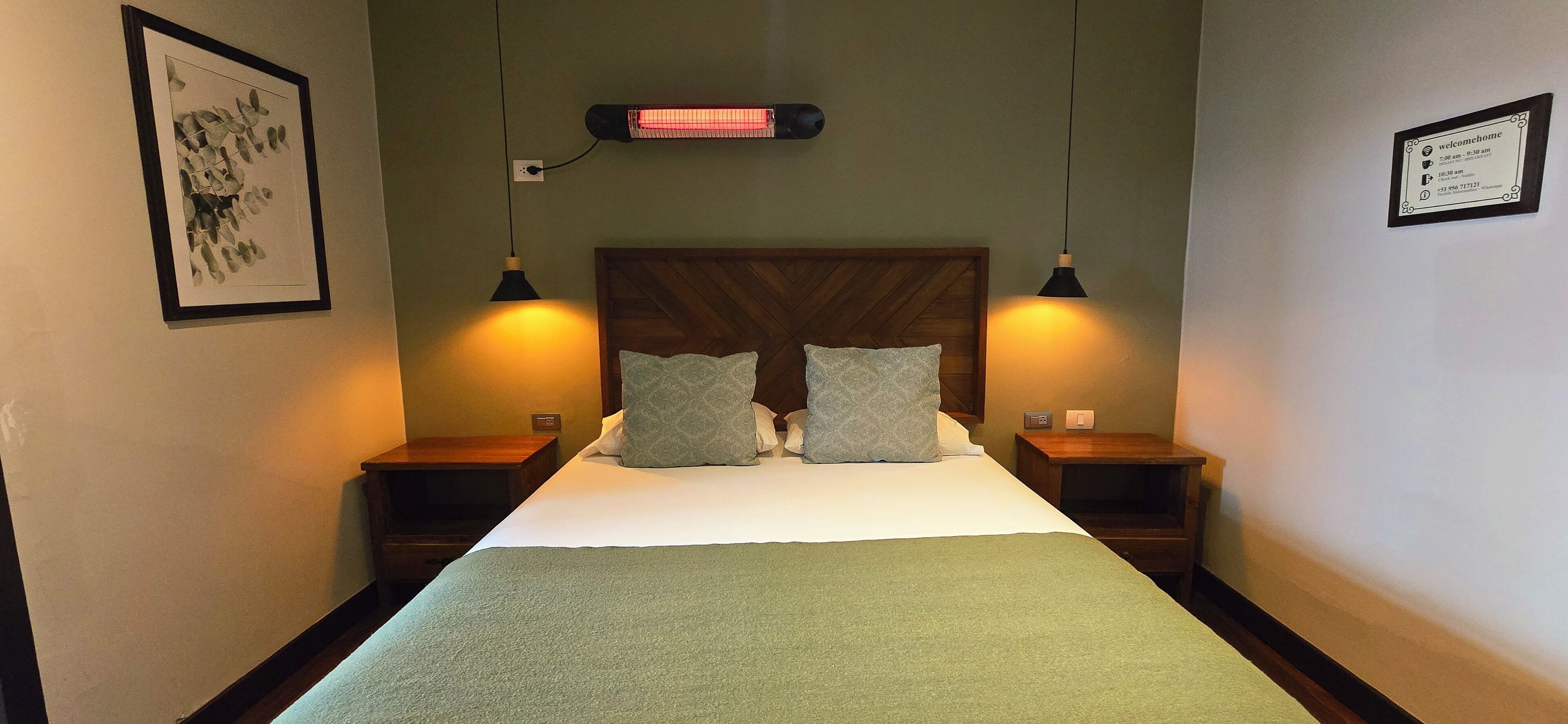 A hotel room with a large bed, two bedside tables, a wall-mounted heater, and a framed welcome sign.