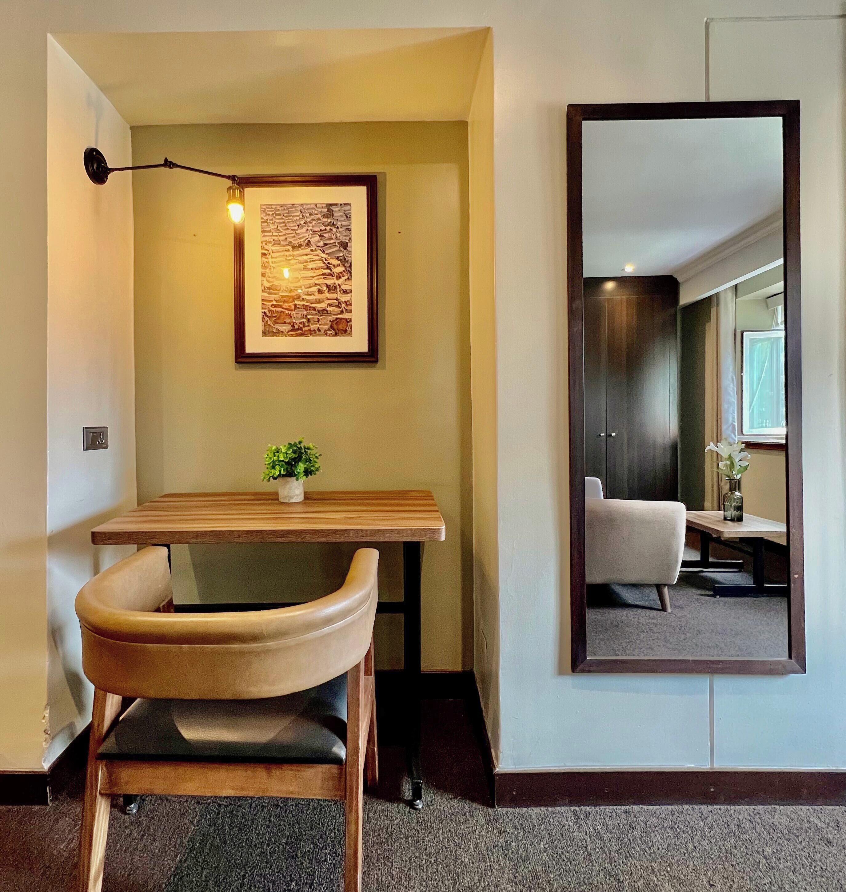 A hotel room with a desk, chair, framed artwork, and a mirror reflecting the room.
