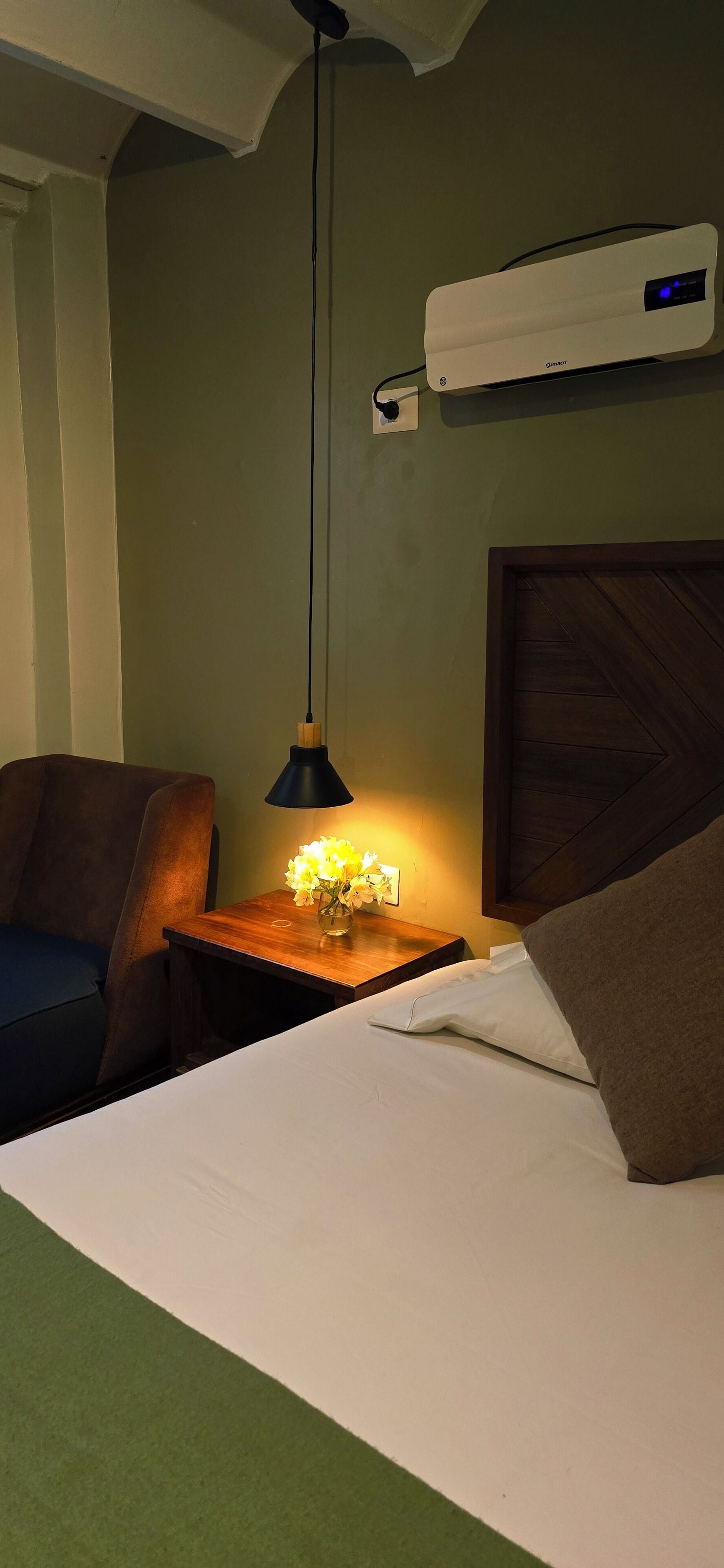 Et hotelværelse med en seng, et natbord med lampe, en vægmonteret aircondition og en træsenggavl.