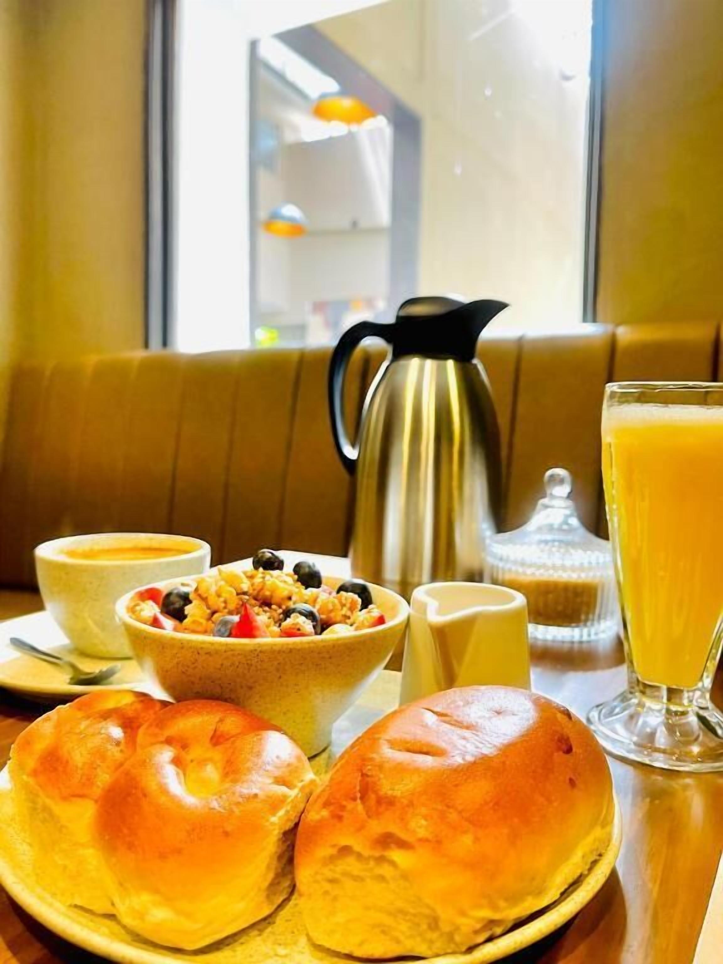 Et morgenbord med skåls havregryn, en kande, et glas appelsinjuice og to rundstykker på et bord.