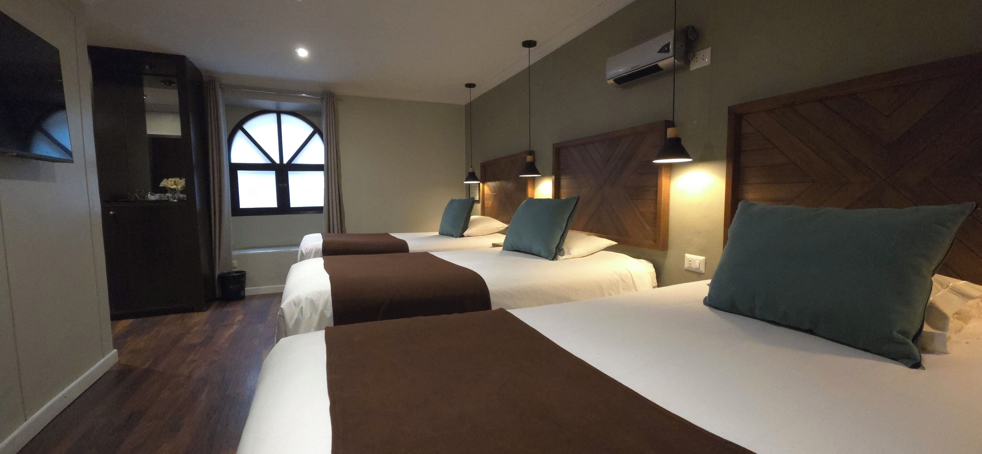 A hotel room with three beds, a wooden headboard, a large window, and a TV on the wall.