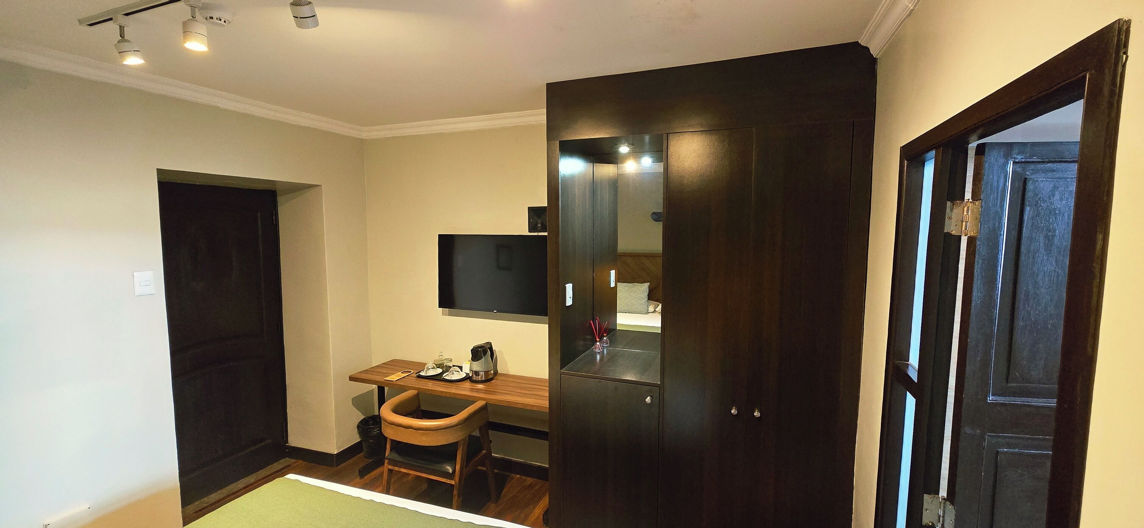A hotel room with a desk, chair, TV, wardrobe, and a small trash bin.