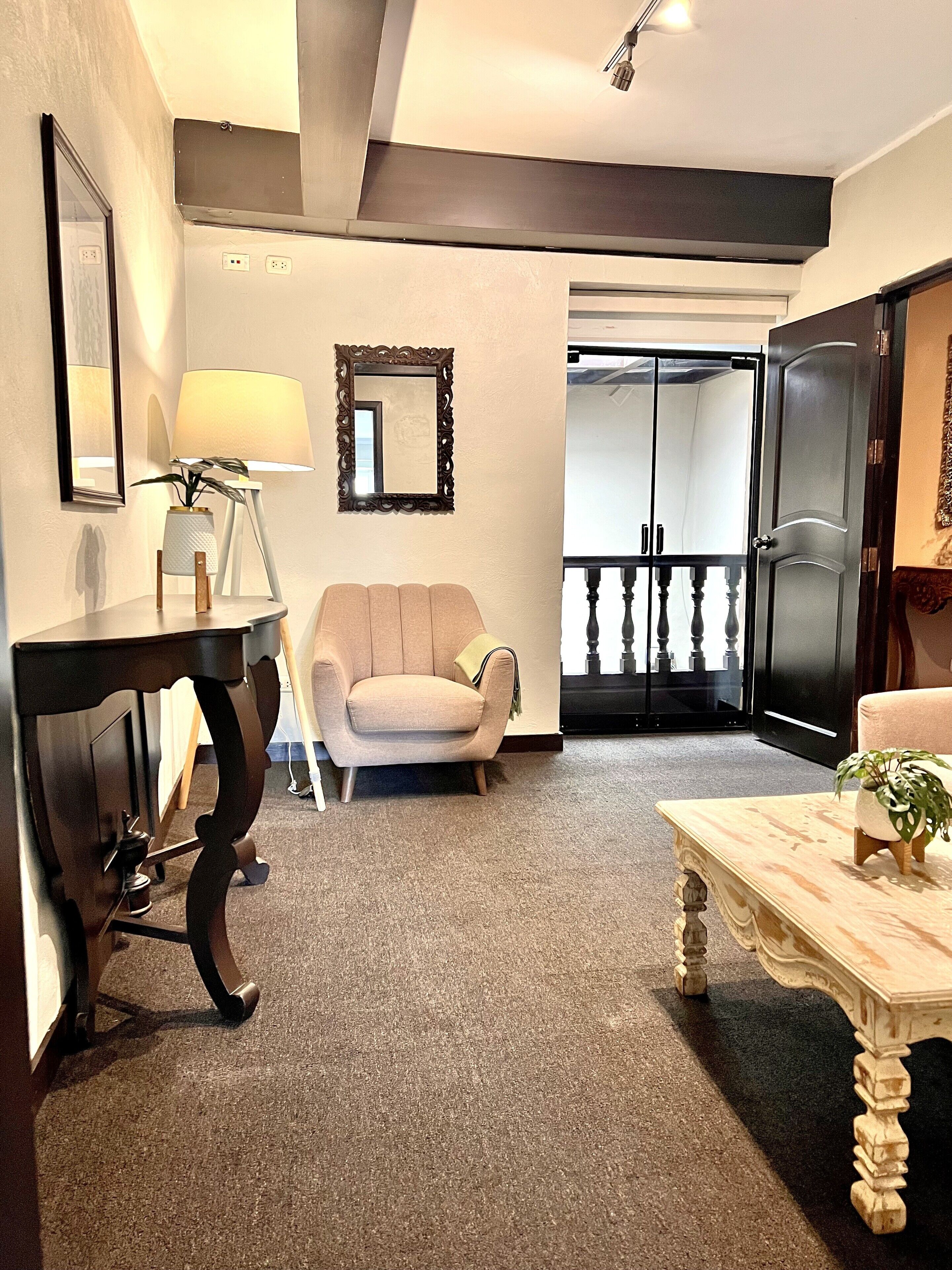 A living room with a sofa, a table, a lamp, and a mirror. There is a door leading to a balcony.