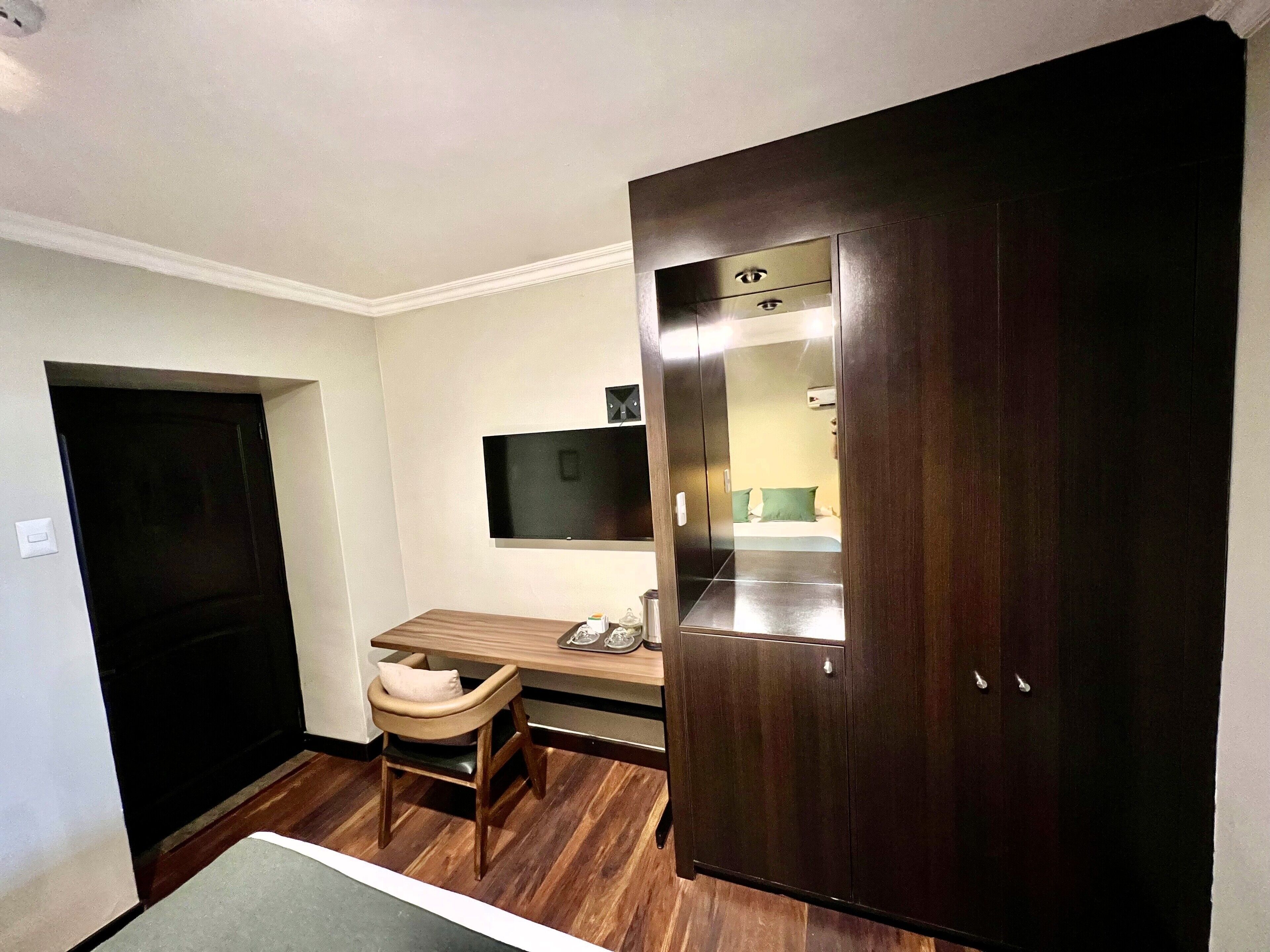 A small room with a wooden desk, a chair, a dark wooden wardrobe, a flat-screen TV mounted on the wall, and a door.