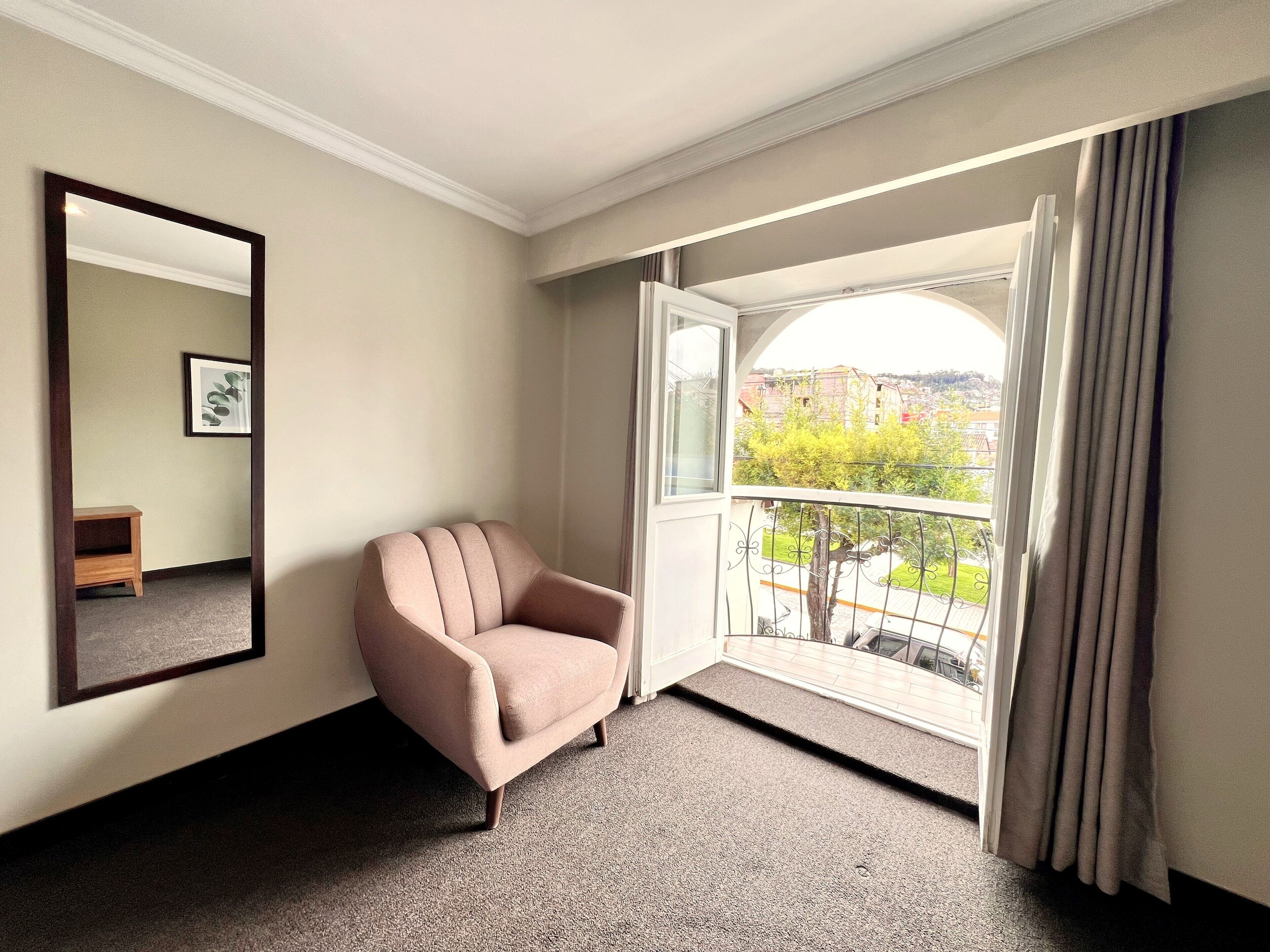 A hotel room with a large window, a chair, a small table, and a mirror.