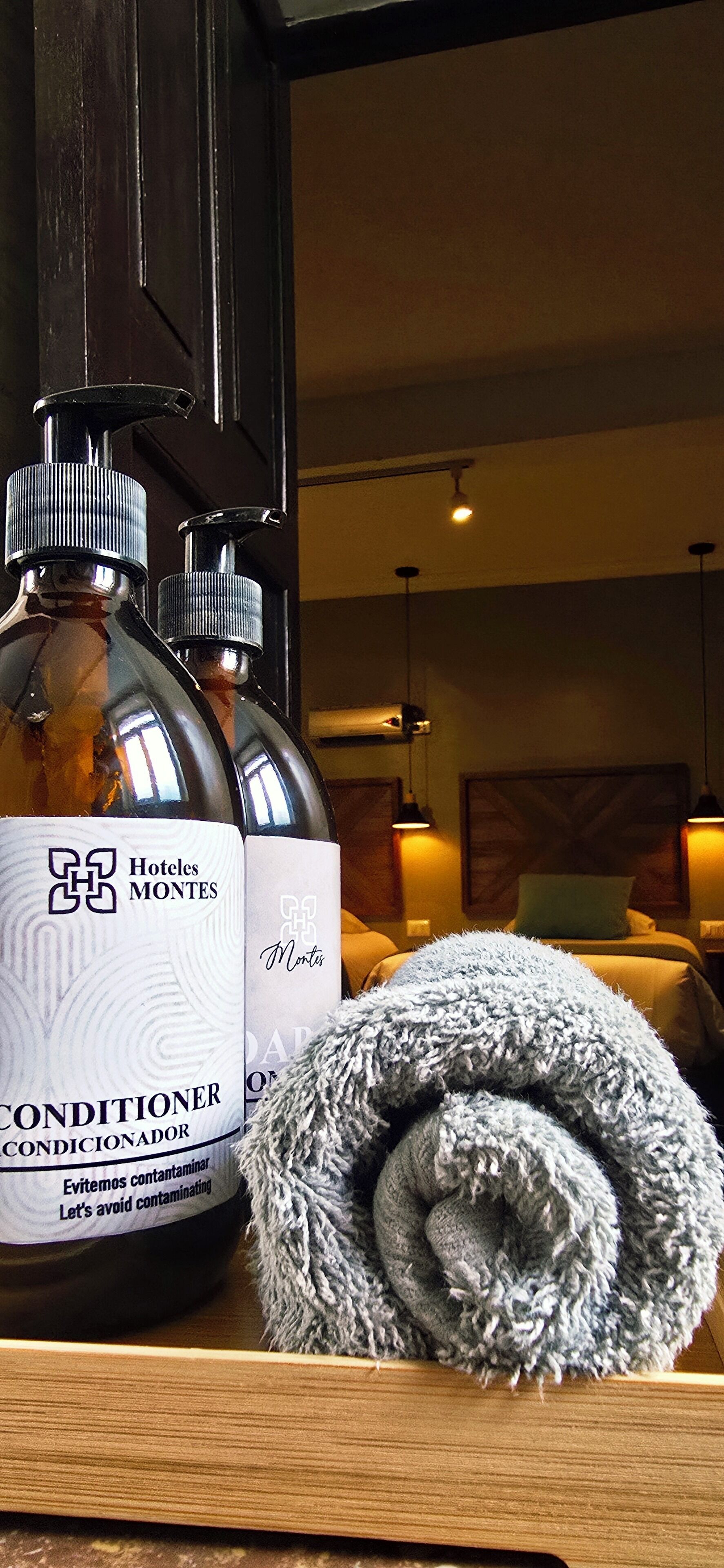 Two bottles of Hotel Montes conditioner and a rolled towel on a wooden surface.
