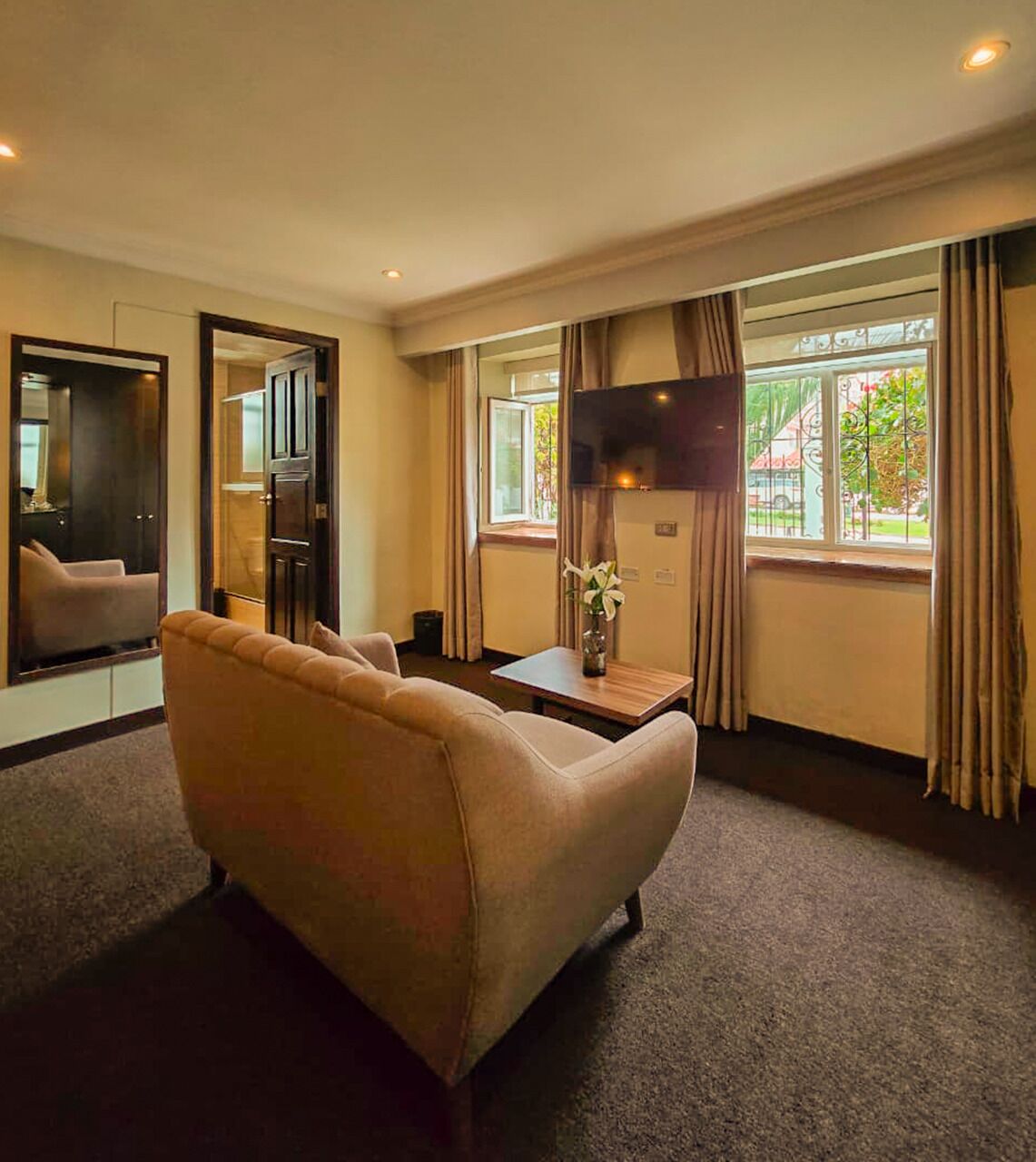 A hotel room with a sofa, a TV, a small table with a vase, and a window with curtains.