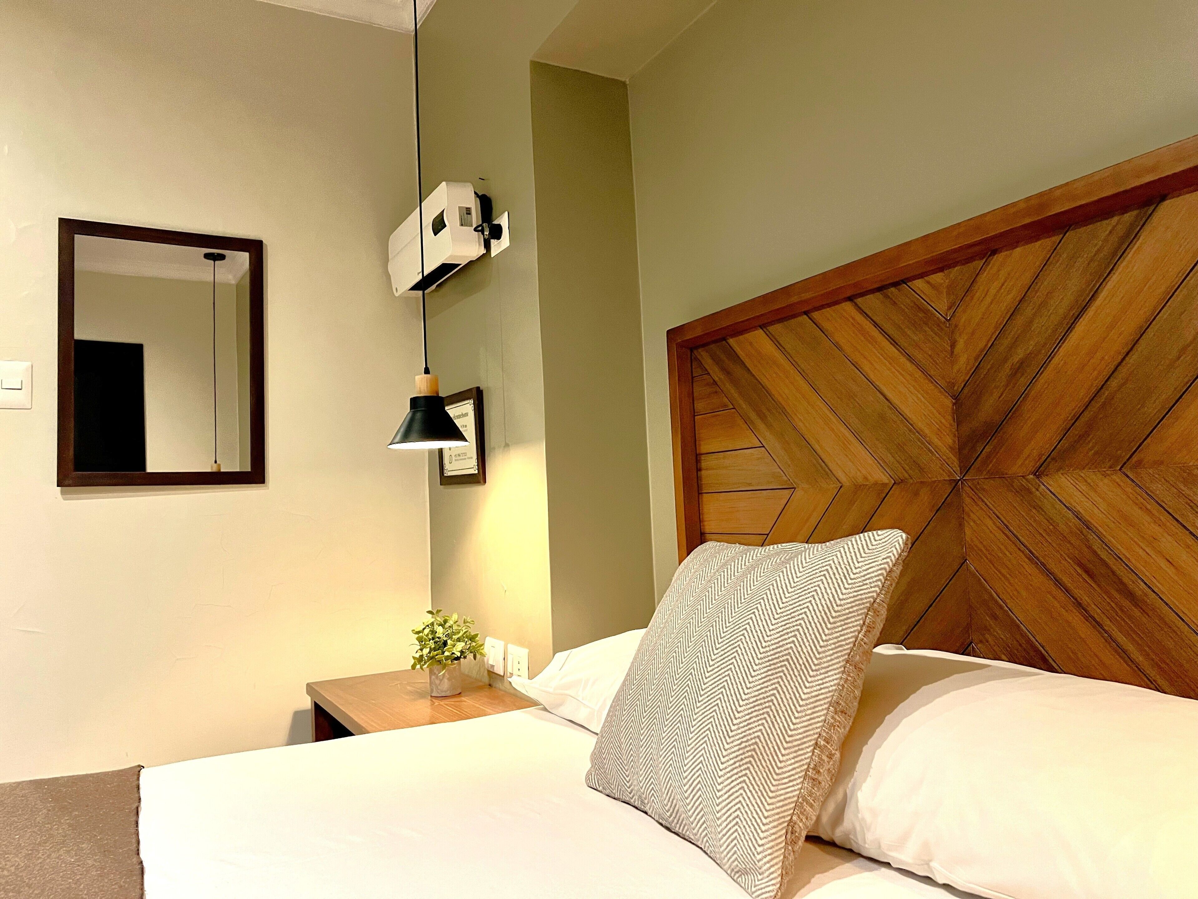 Una cama bien tendida con cabecera de madera, una mesita de noche con lámpara, un espejo y un certificado enmarcado en la pared.