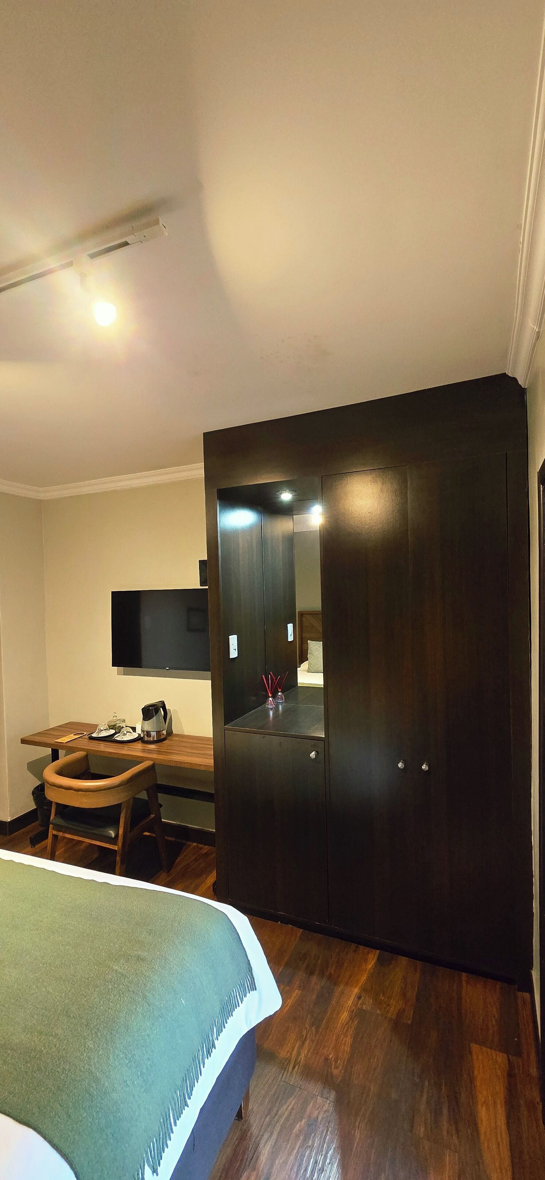 A bedroom with a bed, a desk, a chair, a wardrobe, and a television on the wall.