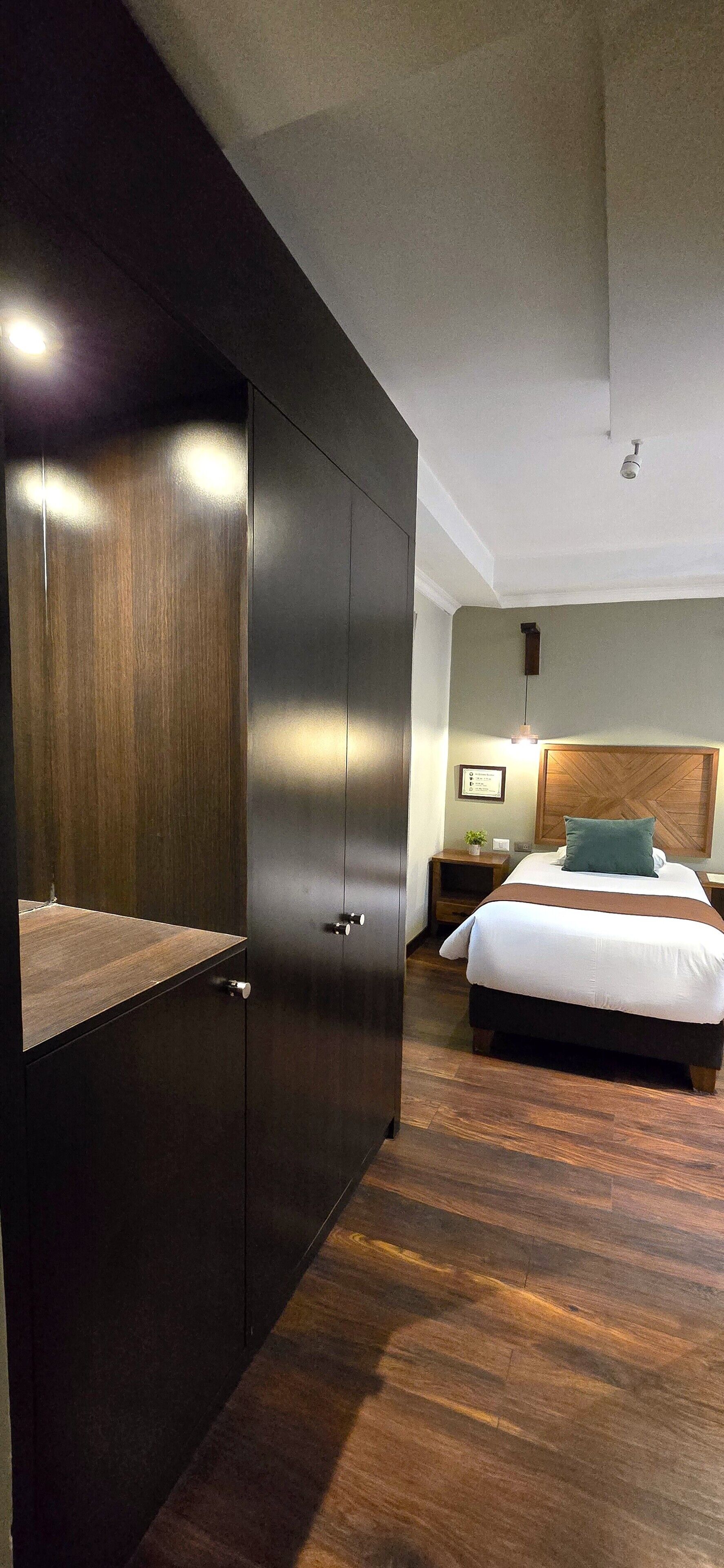 A hotel room with a large wardrobe, a bed with a headboard, a bedside table, and a nightstand.