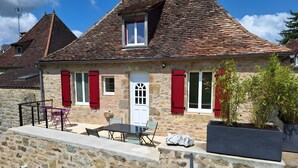 Un bâtiment en pierre avec un toit en tuiles, des volets rouges, une porte blanche et des sièges à l’extérieur.