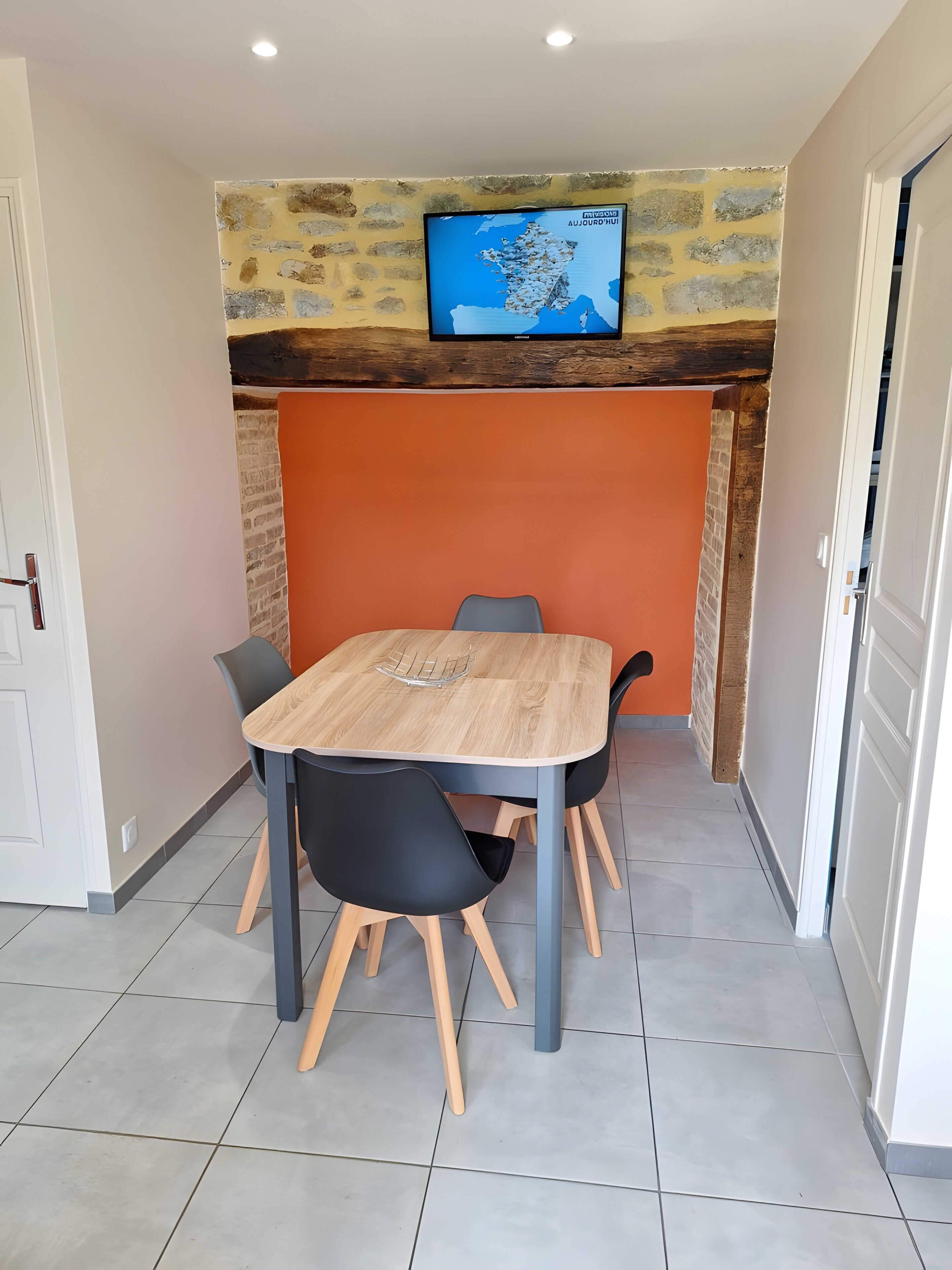 Un coin repas avec une table et des chaises en bois, un téléviseur fixé à un mur en pierre et une porte sur la droite.
