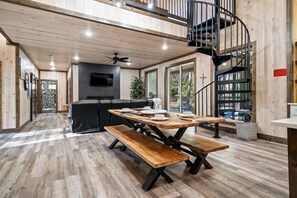 Amplia habitación con suelo de madera, una mesa de comedor con bancos, un sofá negro, un televisor montado en la pared y una escalera de caracol.