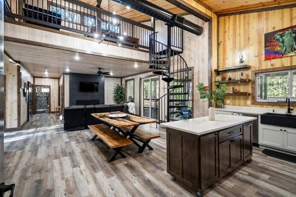 Una cocina amplia con una isla central, un comedor con mesa de madera y una escalera que sube a un nivel superior.