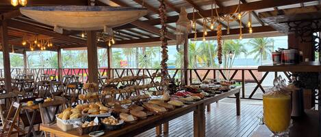 A covered dining area with wooden tables and chairs, a buffet with various food items, and hanging light fixtures.