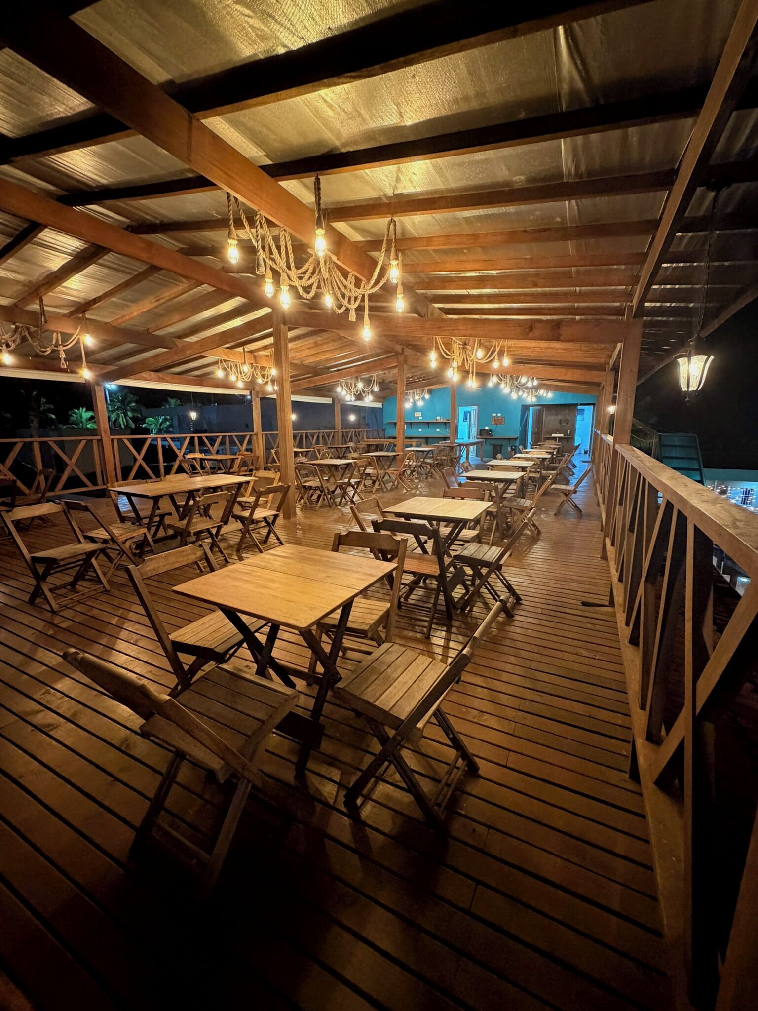 Un espace extérieur aménagé pour les repas, avec des tables et des chaises en bois, un toit abrité et des luminaires suspendus.
