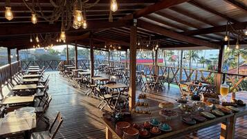 Un restaurante al aire libre con mesas y sillas de madera, un bufé con diferentes platos y vistas al océano.