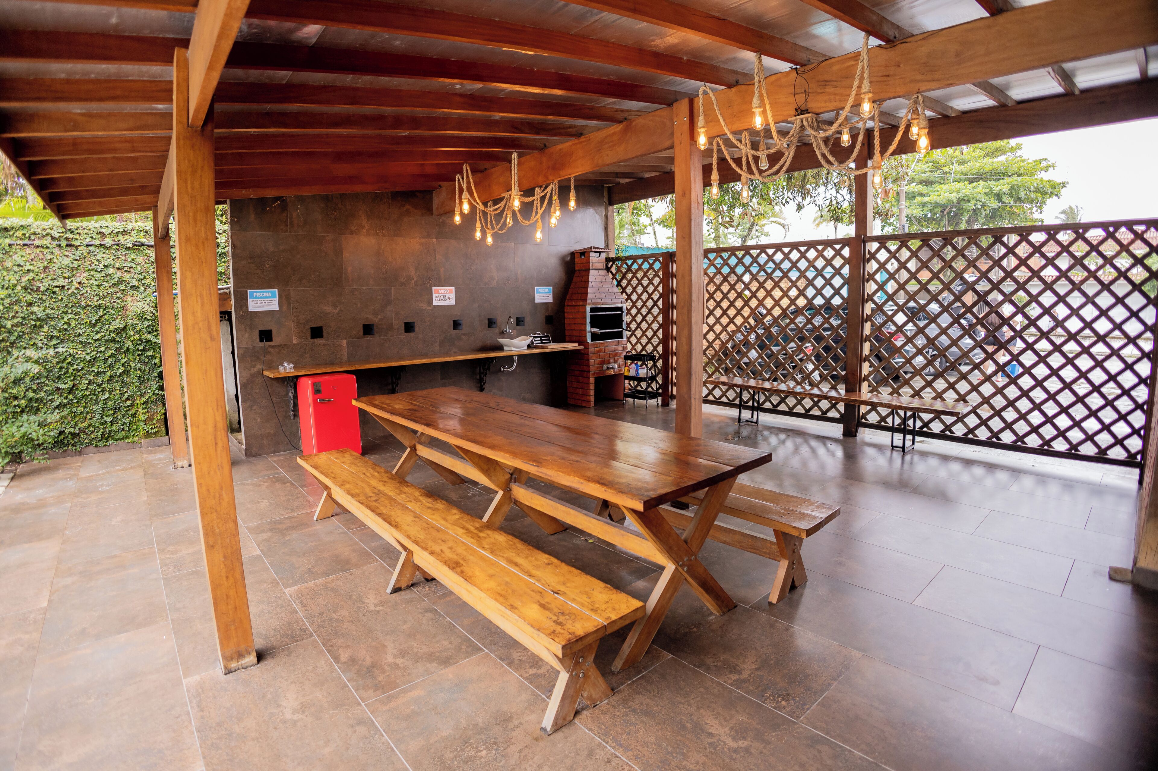 An outdoor patio with wooden tables and benches, a brick oven, and a wooden pergola with string lights.