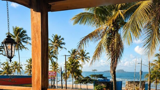 A wooden table set with plates, glasses, and a teapot, overlooking a beach with palm trees and the ocean. - Santa Clara Pousada (Guarujá)