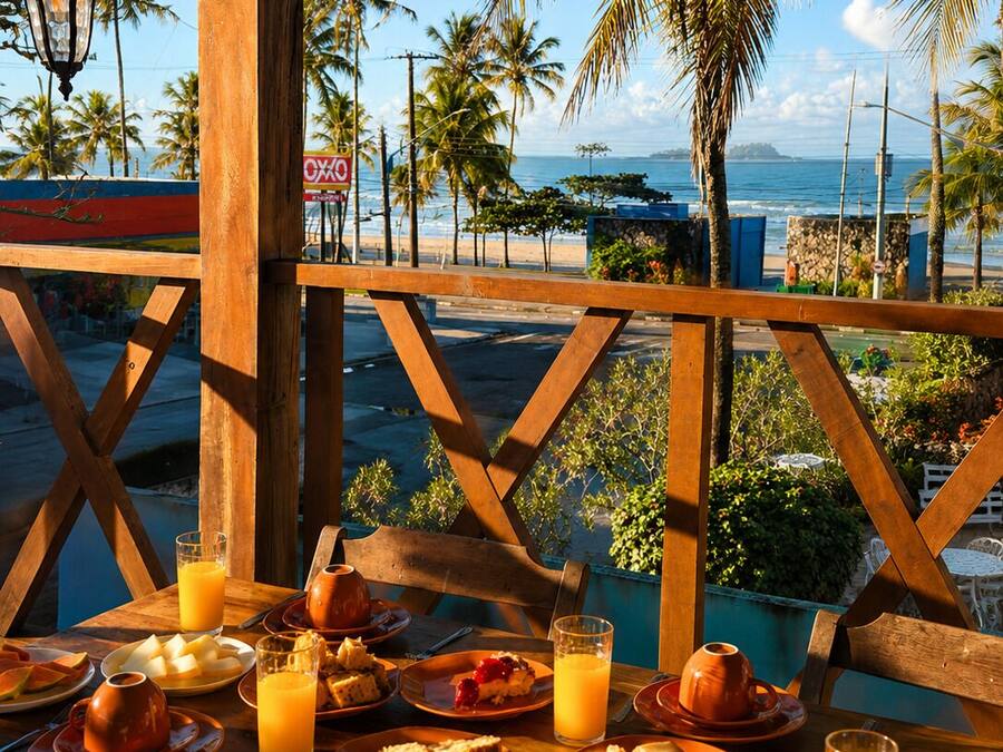 Une table en bois, dressée avec des aliments pour le petit-déjeuner et du jus d’orange, donnant sur une plage bordée de palmiers et offrant une vue sur l’océan.
