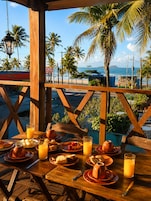Une table en bois, dressée avec des aliments pour le petit-déjeuner et du jus d’orange, donnant sur une plage bordée de palmiers et offrant une vue sur l’océan.