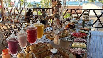 Una mesa de buffet con diferentes alimentos y bebidas, ubicada bajo una estructura de madera con vistas a la playa y palmeras.