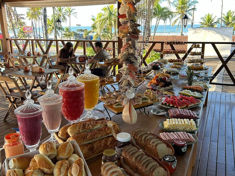 Una mesa de buffet con diferentes alimentos y bebidas, ubicada bajo una estructura de madera con vistas a la playa y palmeras.