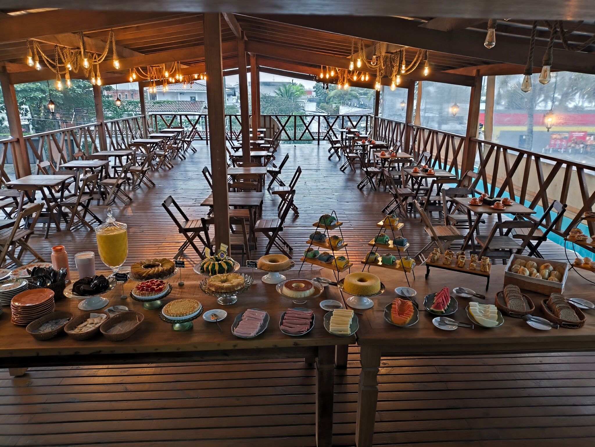 Un área de comedor al aire libre con mesas y sillas de madera, un bufé con diversas opciones de comida y vistas del paisaje circundante.