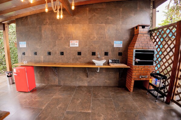 A covered outdoor area with a brick oven, a sink, and a red refrigerator. - Santa Clara Pousada (Guarujá)