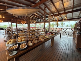 A buffet setup with various food items on a wooden table, under a covered area with hanging lights and a view of the ocean.