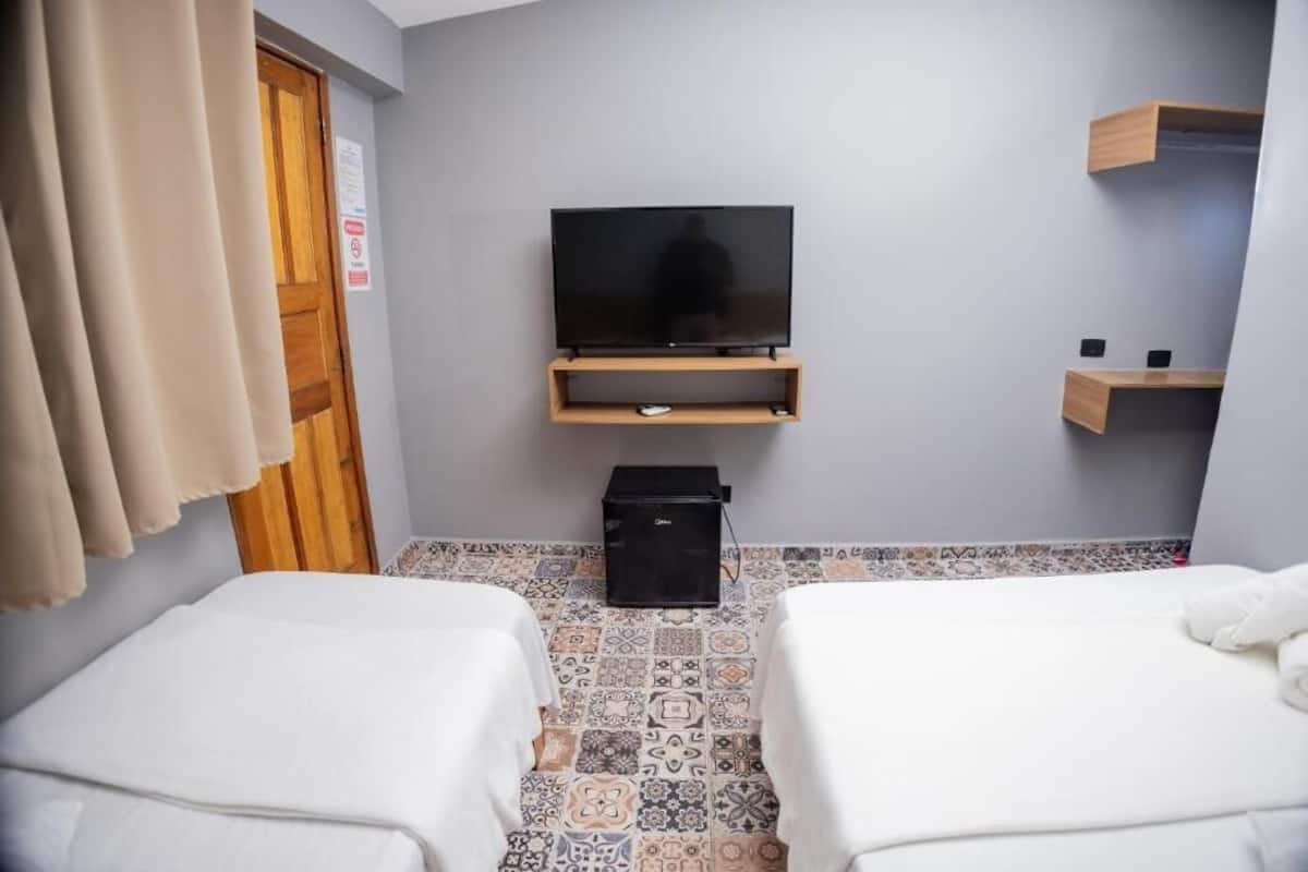 Ein Hotelzimmer mit zwei Betten, einem Fernseher auf einem Holzständer, einem kleinen schwarzen Lautsprecher und einem gefliesten Boden.