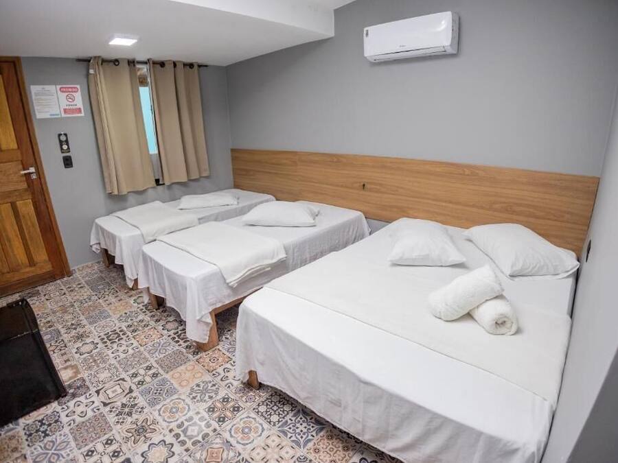 Habitación de hotel con tres camas individuales, cabecera de madera, suelo de baldosas y una ventana con cortinas.