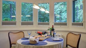 Un coin repas avec une table ronde dressée pour quatre personnes, entourée de chaises en osier. Des fenêtres offrent une vue sur les arbres, et des lampes suspendues illuminent la table.