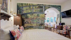 Une chambre avec un grand lit, un tableau représentant un pont, un coin repas avec des chaises et un téléviseur fixé au mur.
