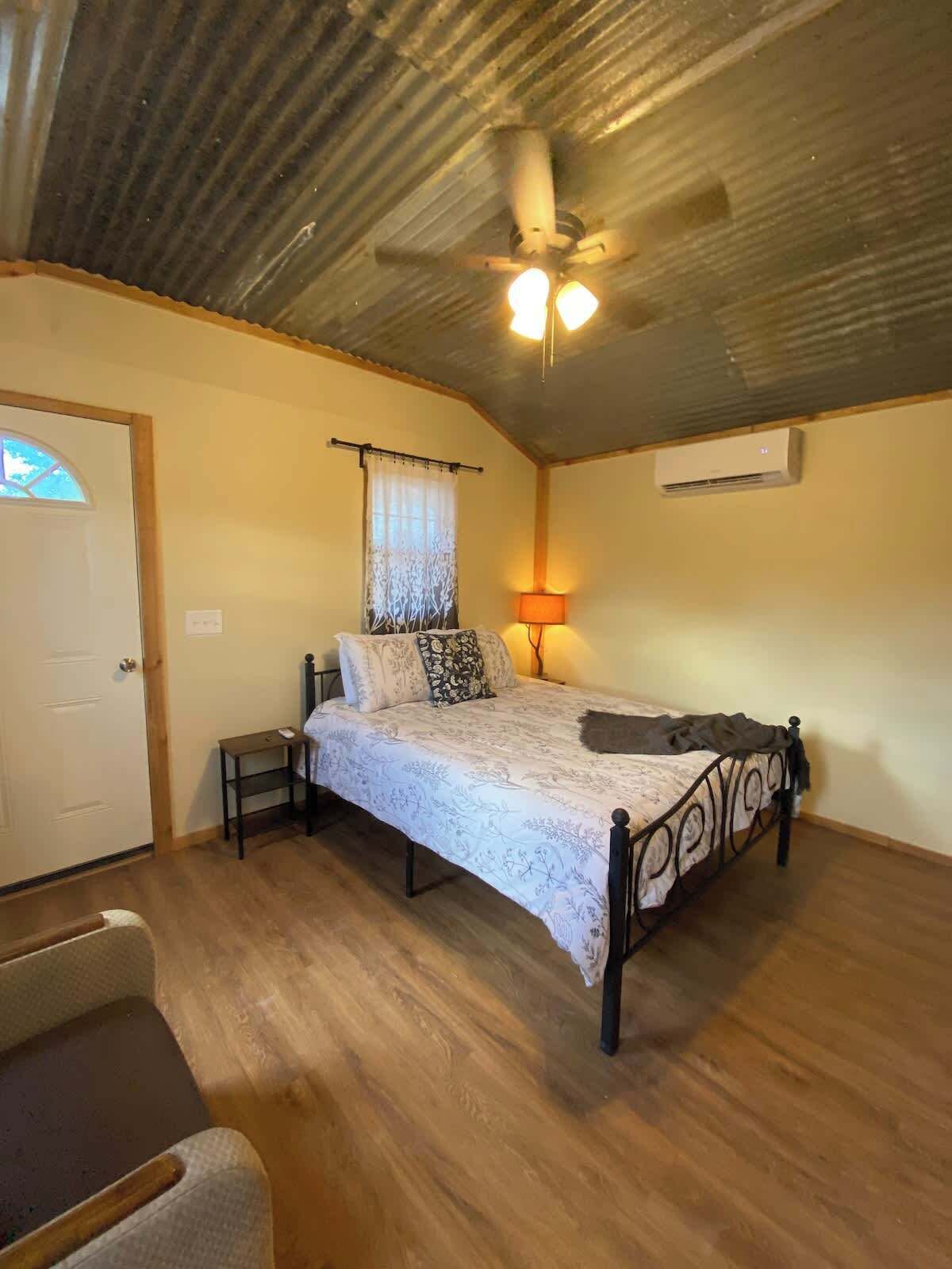 Une chambre à coucher comprenant un lit, un ventilateur de plafond, un appareil de climatisation, une lampe de chevet et une porte.