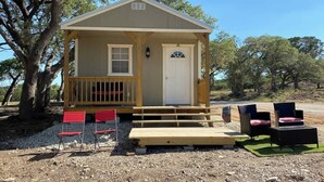 Una cabaña pequeña de un solo piso, con un porche, una puerta principal y una ventana. Hay mobiliario exterior con sillas y una mesa.