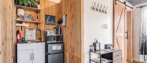 Et kompakt køkken med trævægge, en hvid bordplade og et sort køleskab. Hylder med planter og køkkenudstyr, en hængende lampe og en skydedør i landstil.