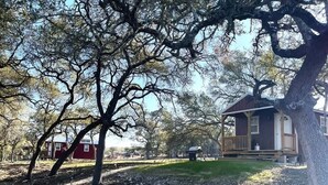 A rustic cabin with a porch, surrounded by trees and a dirt path. - Hill Country Cabin 4 | Cozy Couples Retreat (Canyon Lake)