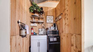 A compact kitchen with wooden walls, white cabinets, a black refrigerator, a microwave, and a coffee maker. - Hill Country Cabin 4 | Cozy Couples Retreat (Canyon Lake)