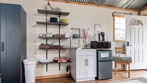 A compact kitchen with a refrigerator, microwave, and shelves with various items. - Hill Country Cabin 3 | Sleeps 4, Gas Grill (Canyon Lake)