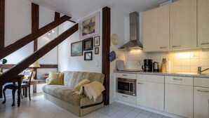 A cozy living room with a sofa and a dining area, a kitchen with white cabinets and a stove, and a staircase with exposed wooden beams. - Strandvilla Hubertus - H6 - Paul Signac (Ostseebad Boltenhagen)