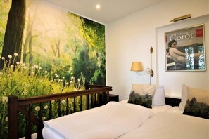 Een slaapkamer met een grote muurschildering van een bos, een bed met witte beddengoed, een nachtkastje, een lamp en een poster aan de muur.